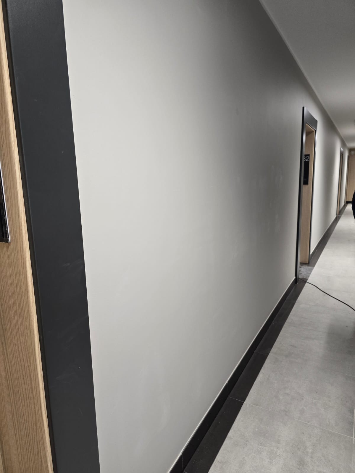 Korytarz po remoncie: gładkie, jasne ściany, ciemne listwy przypodłogowe i obramowania drzwi. Widoczne drzwi z numerem '82'.