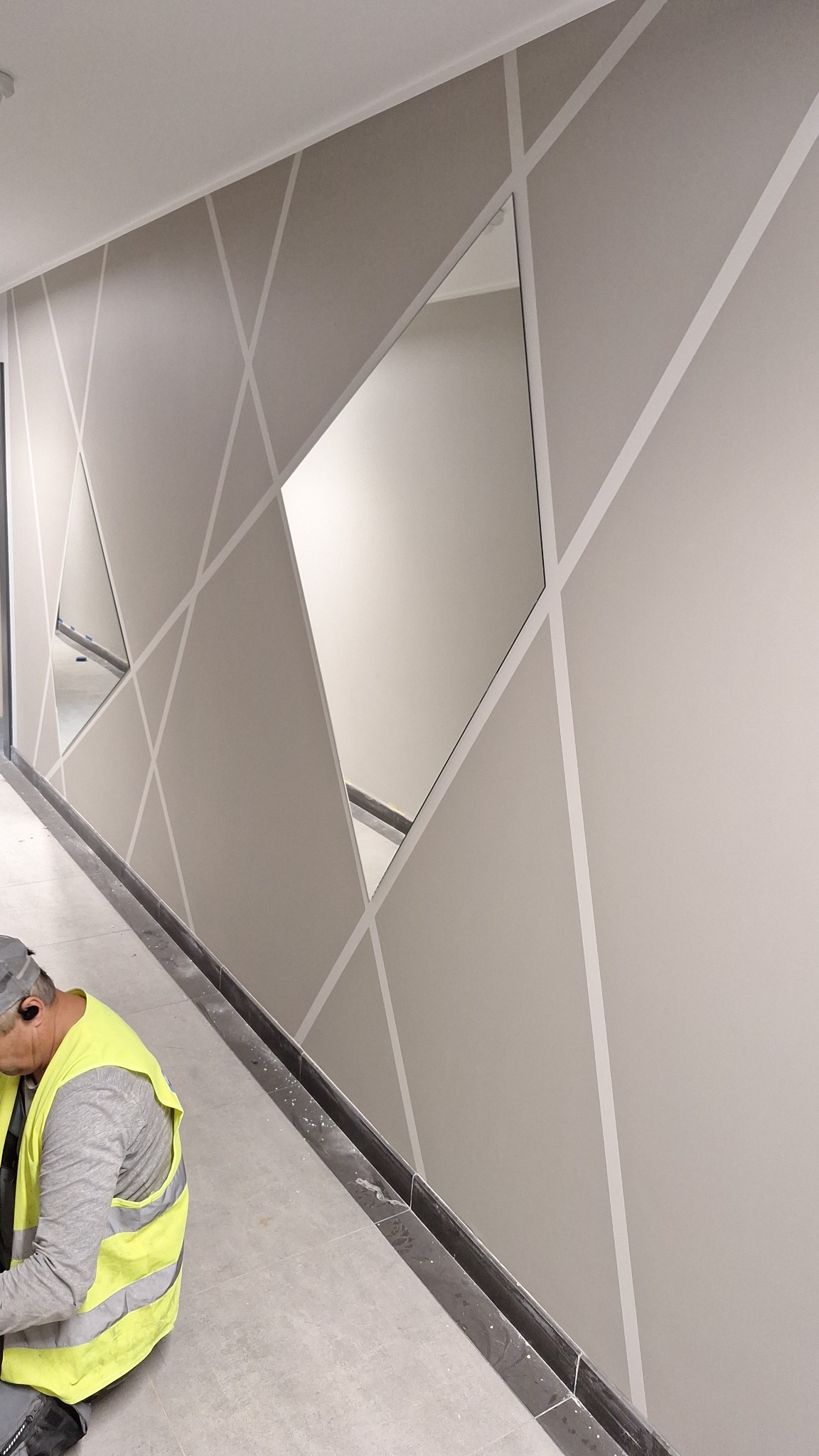 Ściana z geometrycznym wzorem i wkomponowanymi lustrami, pracownik w kamizelce odblaskowej w trakcie pracy. Wykończenie wnętrz w nowoczesnym stylu.