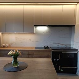 Meble na wymiar - Karol Światłoń - Beżowe szafki kuchenne podświetlone LED z blatem imitującym drewno, marmurowe płytki na ścianie i czarny piec wolnostojący, wazon z kwiatami na drewnianym stole.