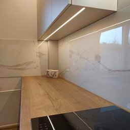 Meble na wymiar - Karol Światłoń - Nowoczesna kuchnia: białe szafki z oświetleniem LED pod spodem, blat drewniany z wbudowaną płytą indukcyjną, marmurowe płytki na ścianie, dwie filiżanki z kwiatowym wzorem w tle.