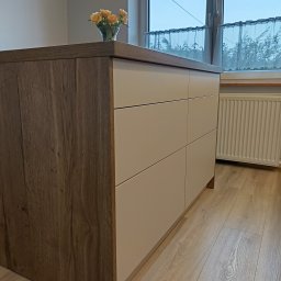 Meble na wymiar - Karol Światłoń - Fragment szafki kuchennej z trzema poziomymi szufladami w kolorze beżowym, obok okna z firanką i wazonem z żółtymi różami, widoczne panele podłogowe.