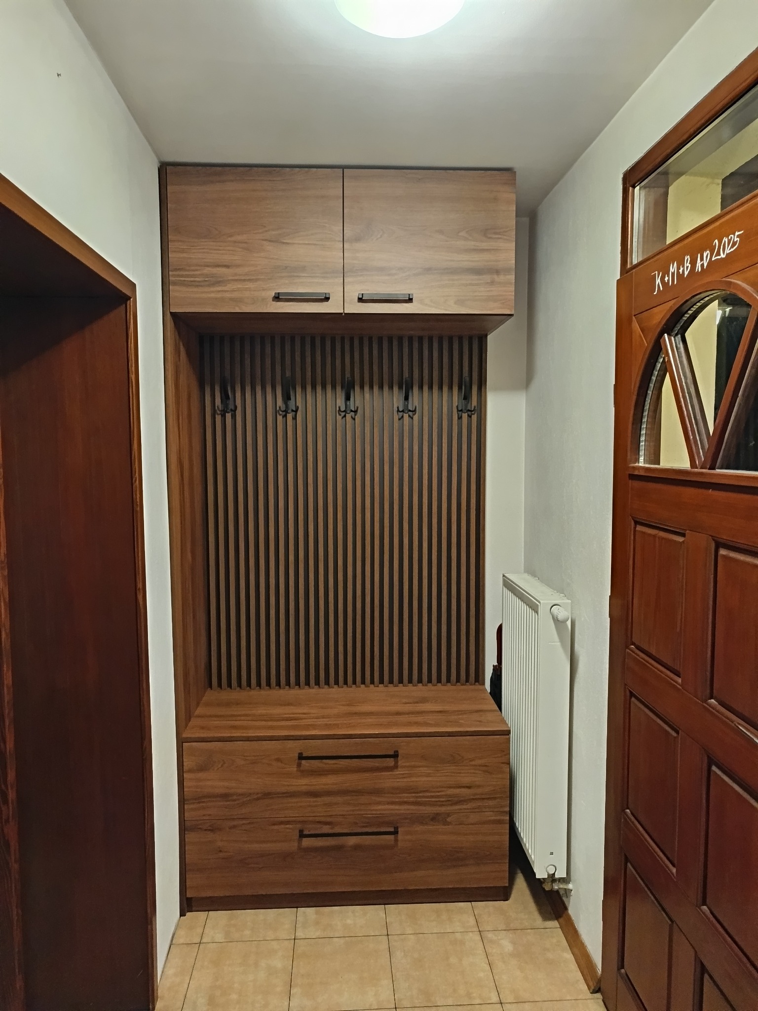 Zabudowa przedpokoju z drewnianymi szafkami górnymi, siedziskiem z szufladami i panelem z wieszakami na ubrania, widoczny grzejnik i drzwi wejściowe z napisem K+M+B AP 2025.
