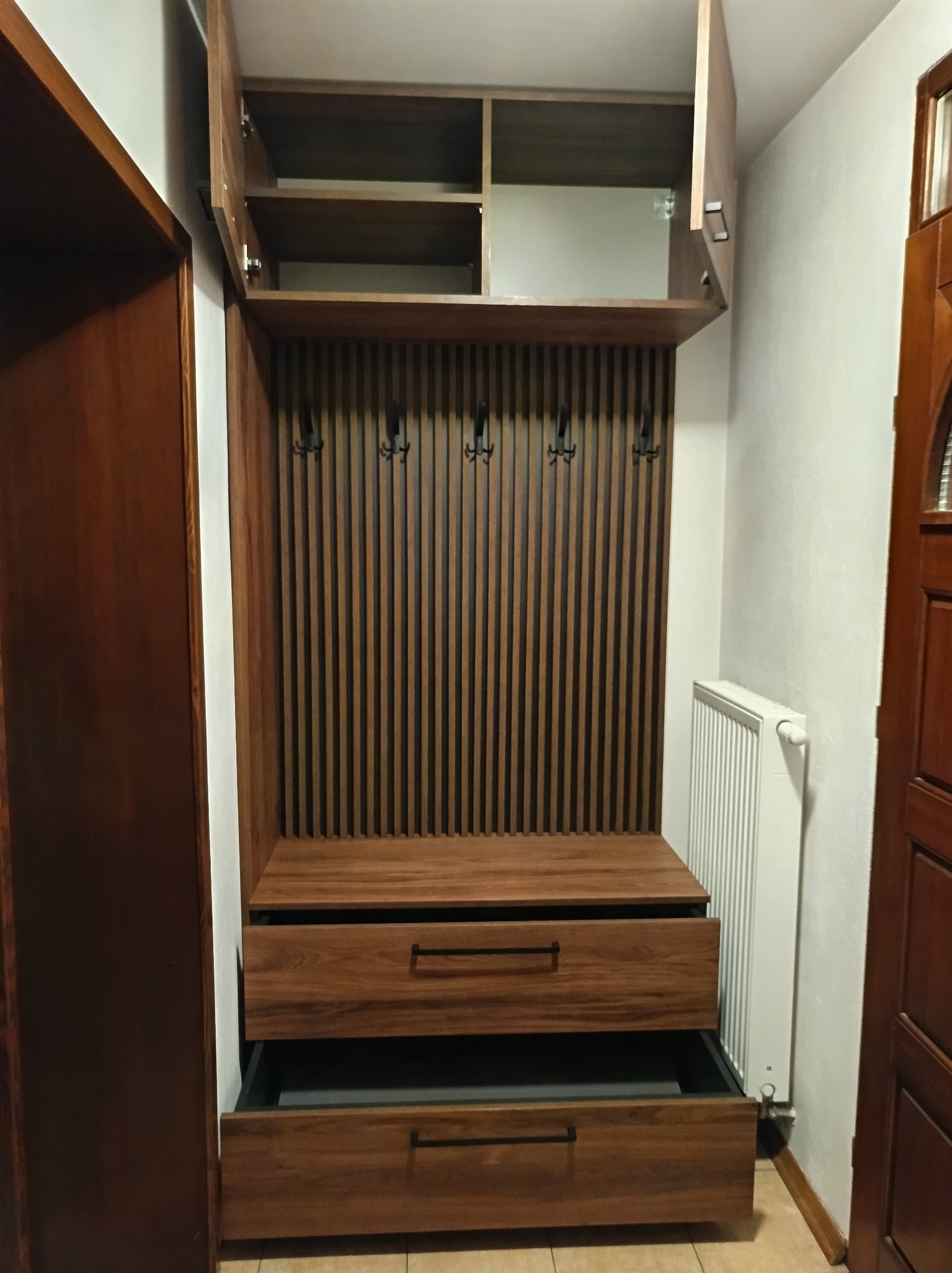 Drewniana szafa w przedpokoju z otwartymi szafkami górnymi i szufladą, w tle grzejnik.