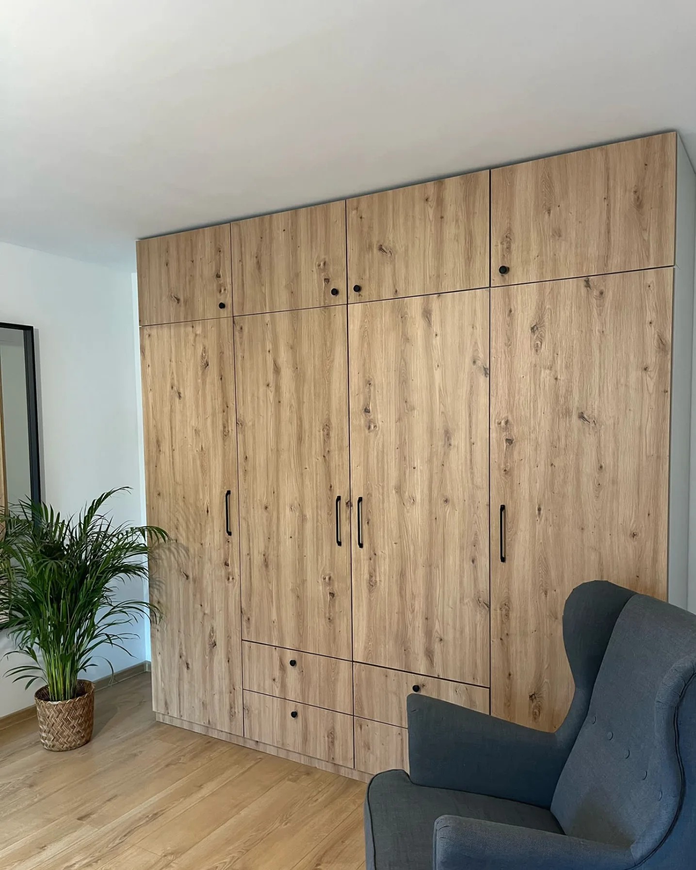 Wysoka szafa wnękowa z drewnopodobnymi frontami, czarne podłużne uchwyty, uzupełniona o nadstawki pod sufit, obok stoi szary fotel typu uszak i roślina doniczkowa.