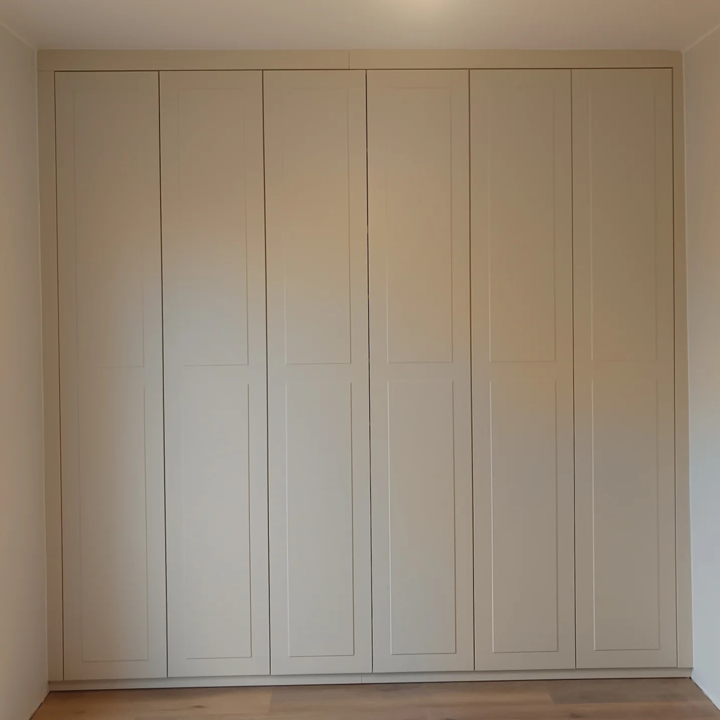 Duża, beżowa szafa wnękowa z sześcioma drzwiami, widoczna na tle jasnej ściany i drewnianej podłogi.