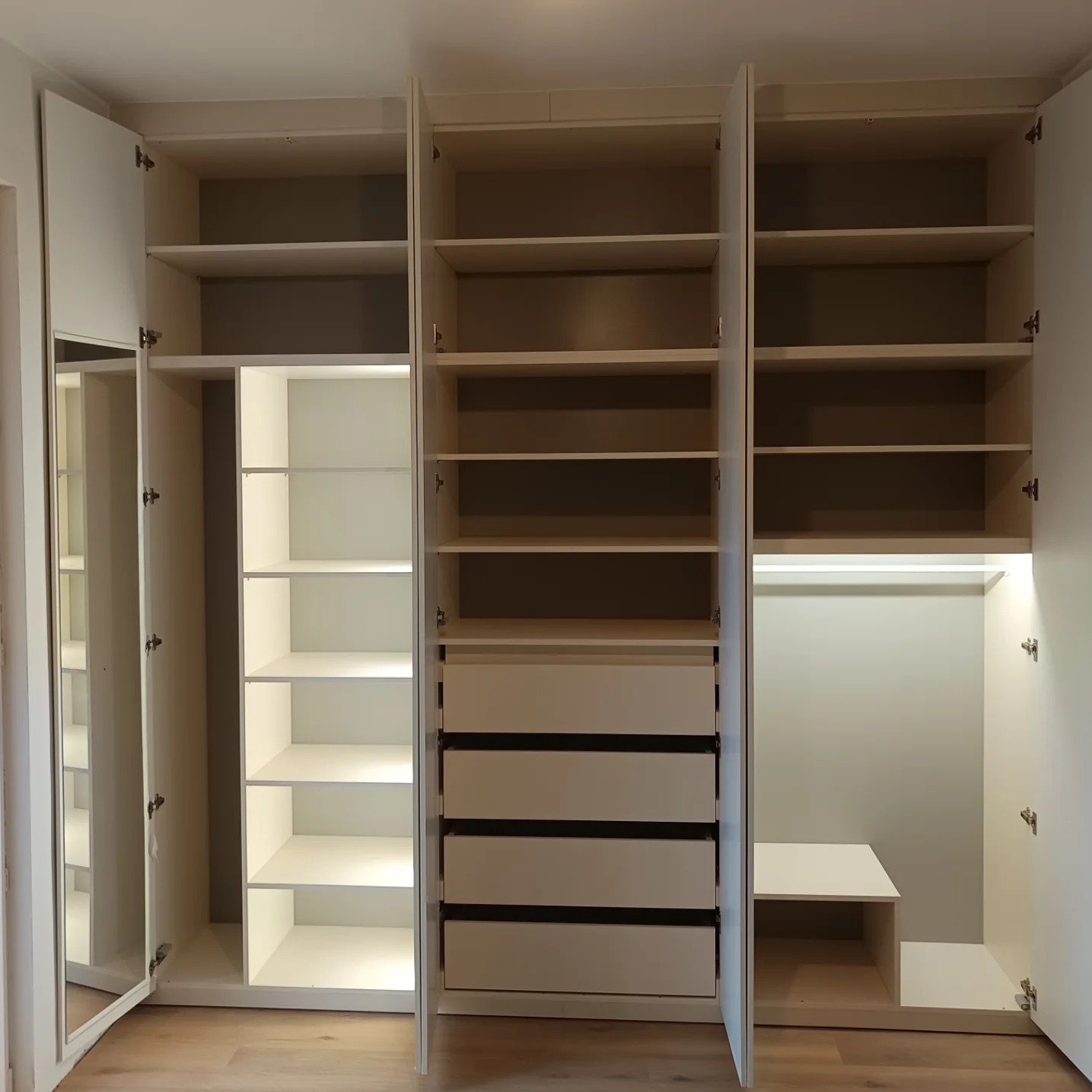 Otwarta, biała szafa wnękowa z lustrzanymi drzwiami, półkami i szufladami, oświetlona od wewnątrz, na jasnej podłodze.