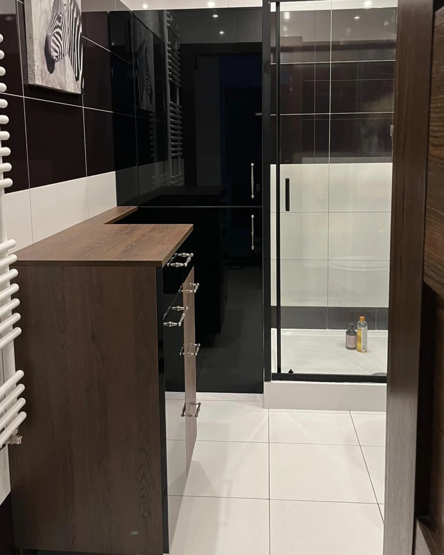 Nowoczesna łazienka z ciemnymi płytkami, szafką pod umywalkę z drewnianym blatem i kabiną prysznicową z czarnymi ramami.