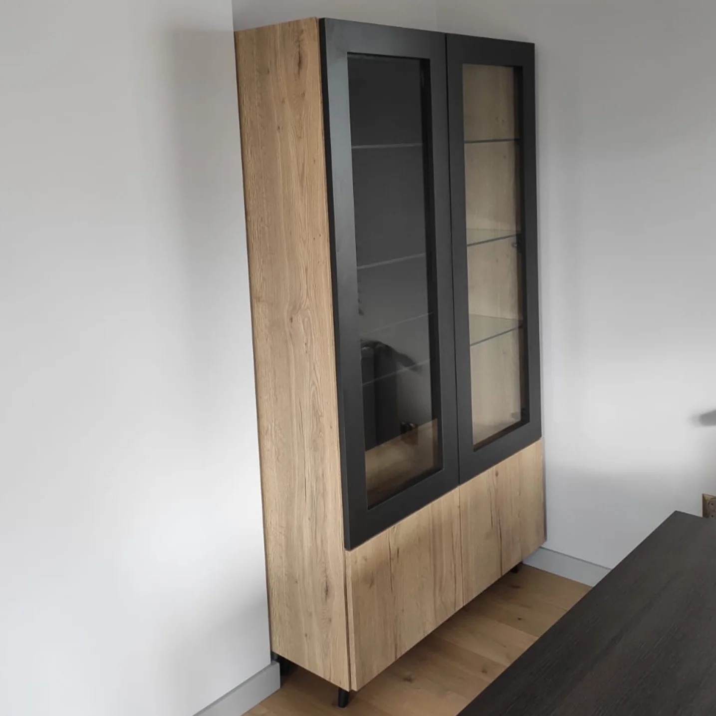 Wysoka szafa z drewnianymi frontami i czarnymi ramami szklanych witryn, na nóżkach, stojąca przy białej ścianie.