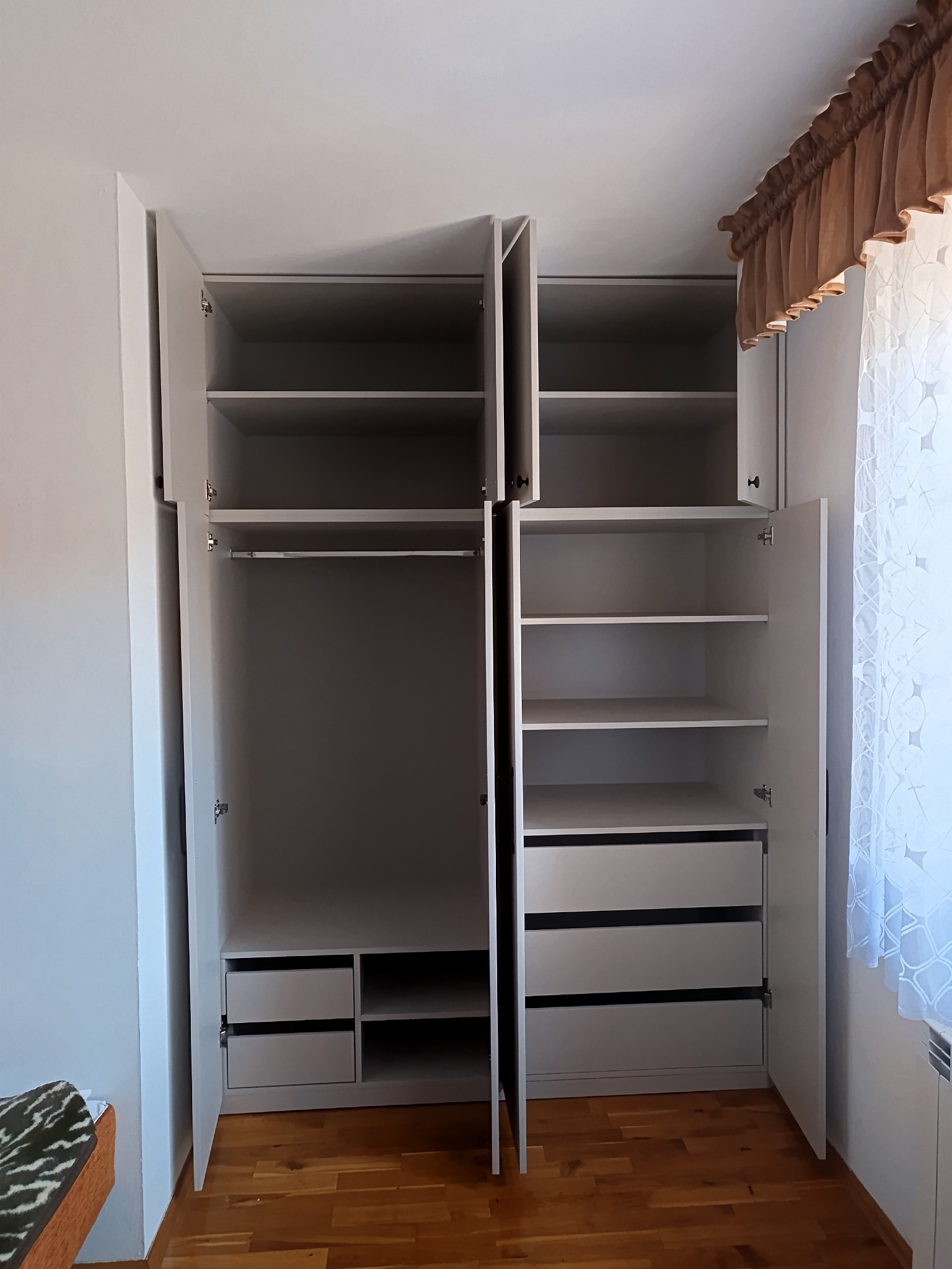 Wnętrze białej, narożnej szafy wnękowej z otwartymi drzwiami, ukazującą półki, szuflady i drążek na ubrania, na tle drewnianej podłogi i okna z zasłoną.