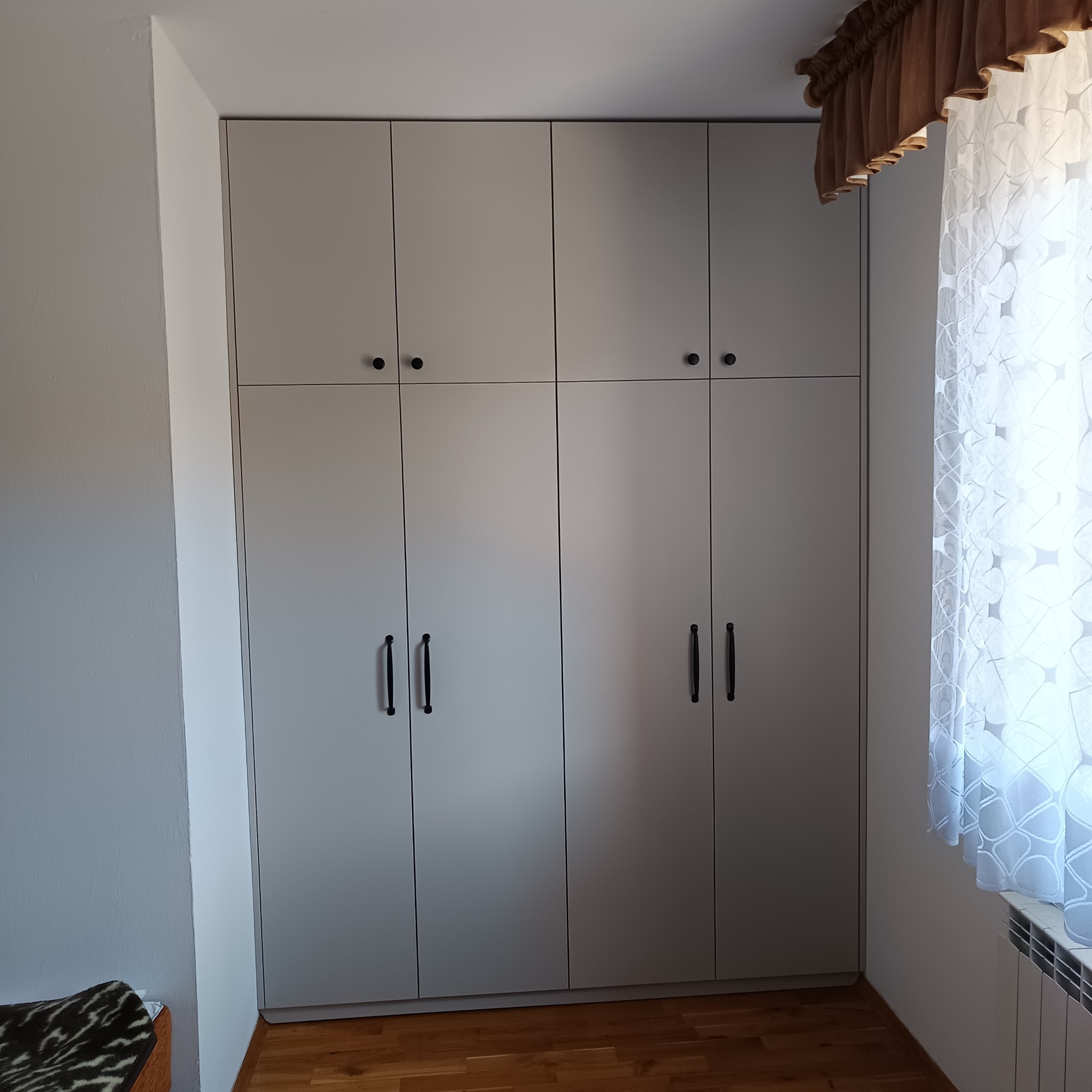 Szara szafa wnękowa z czarnymi uchwytami, idealnie dopasowana do wnętrza w Budzowie, widoczna obok okna z firanką i grzejnikiem.