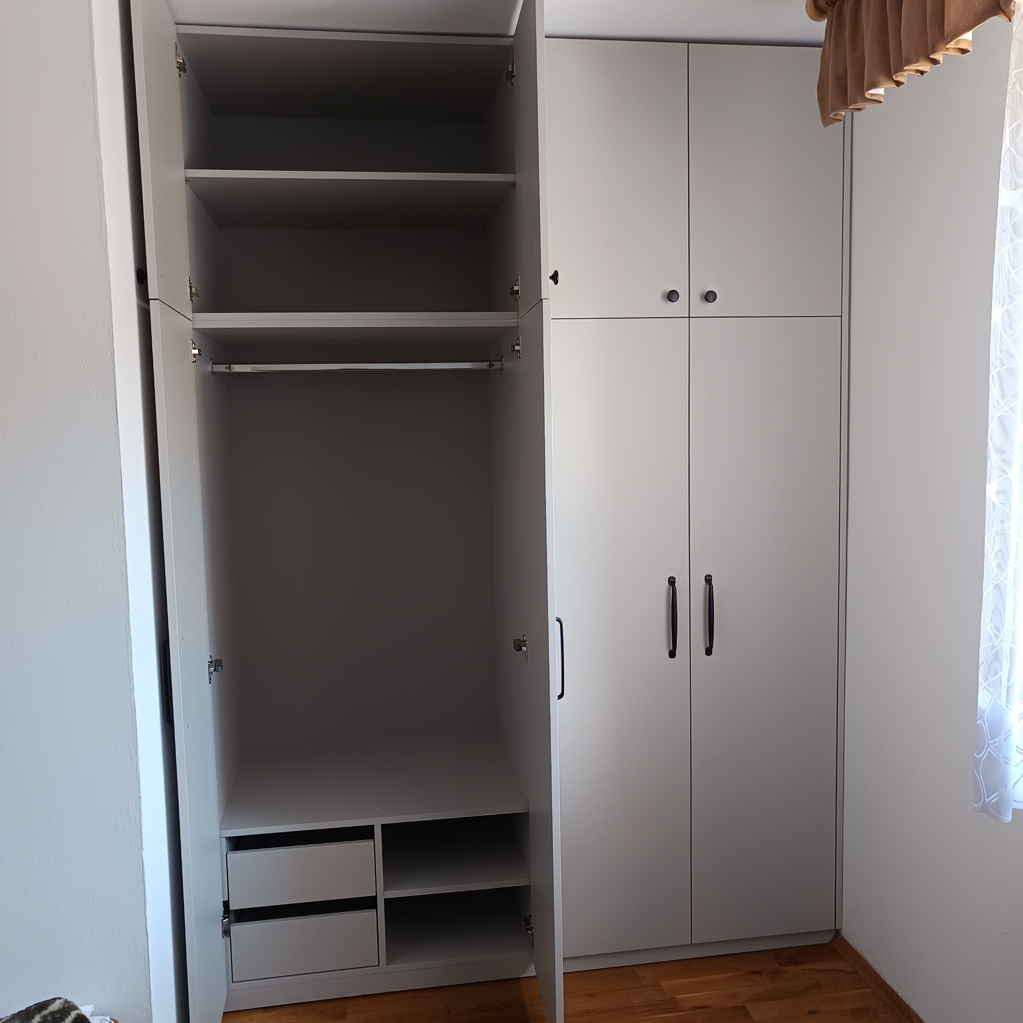 Biała szafa na wymiar z otwartymi drzwiami, widoczne półki, szuflady i drążek na ubrania, czarne uchwyty, drewniana podłoga.