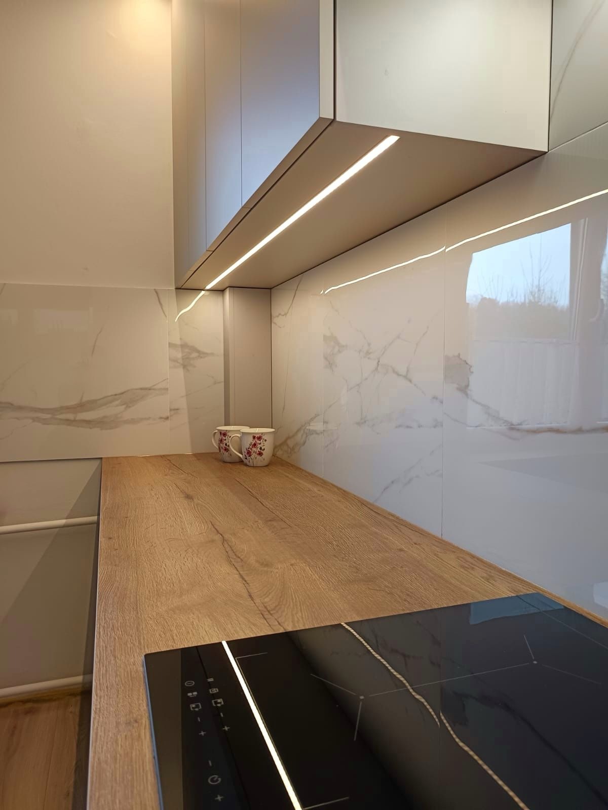 Nowoczesna kuchnia: białe szafki z oświetleniem LED pod spodem, blat drewniany z wbudowaną płytą indukcyjną, marmurowe płytki na ścianie, dwie filiżanki z kwiatowym wzorem w tle.