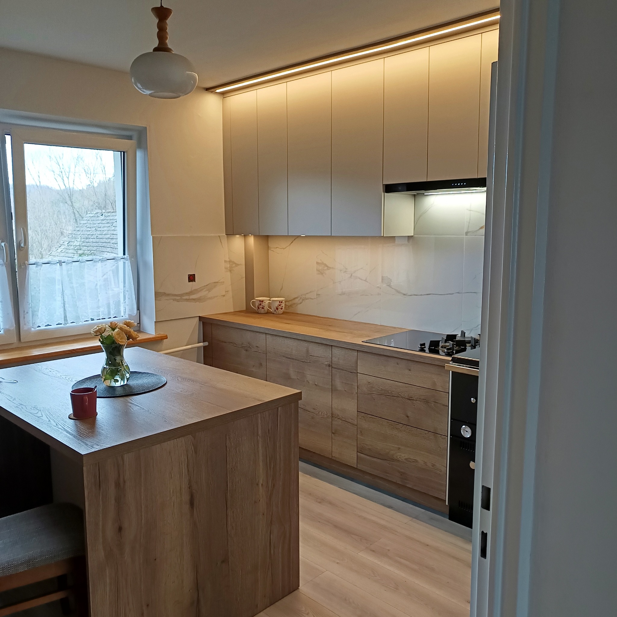 Jasna kuchnia z drewnianymi frontami i wyspą, oświetlenie LED podszafkowe, marmurowy backsplash, widok zza drzwi, bukiet róż na stole.