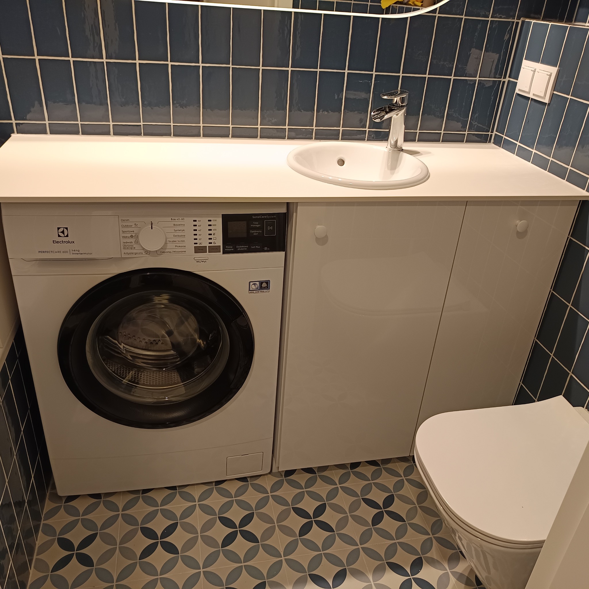 Nowoczesna łazienka z zabudową meblową pod umywalką, z pralką Electrolux PerfectCare 600 wkomponowaną obok szafki, niebieskie płytki na ścianach i wzorzysta podłoga.