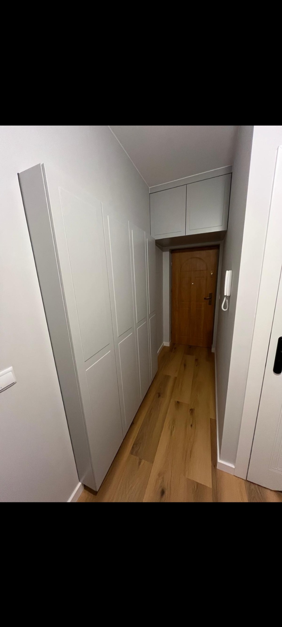 Szara szafa wnękowa z górnymi szafkami w przedpokoju, drewniana podłoga, widok na drzwi wejściowe.