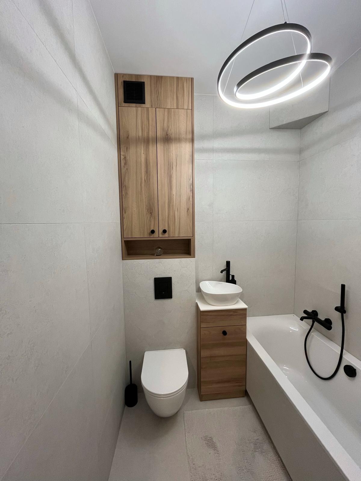 Nowoczesna łazienka z drewnianą szafką wiszącą i umywalką nablatową, czarna armatura, minimalistyczna lampa sufitowa i biała wanna.