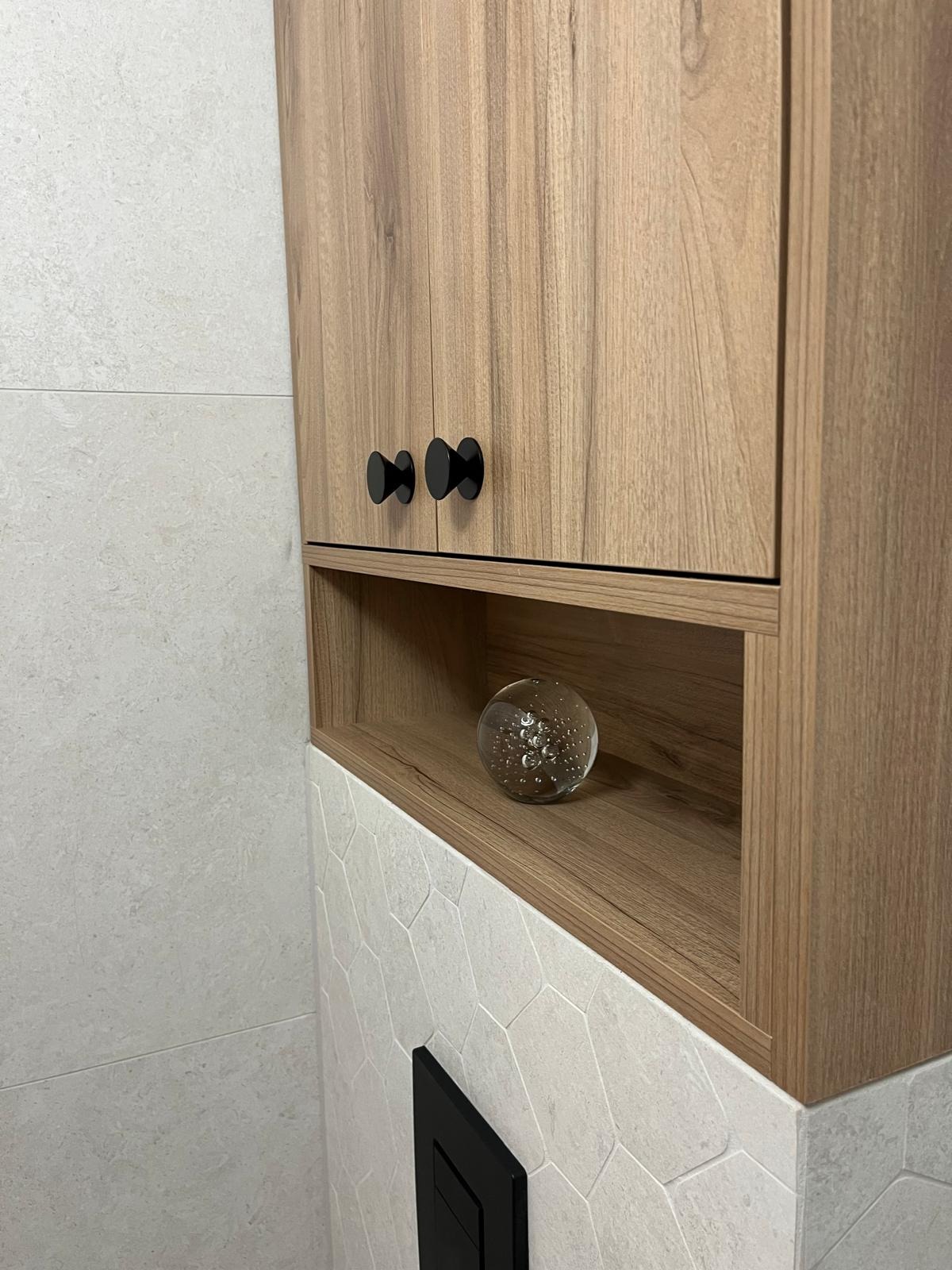 Wisząca szafka łazienkowa z drewnopodobnymi frontami i czarnymi uchwytami, na półce dekoracyjna szklana kula, tło z jasnych płytek heksagonalnych.