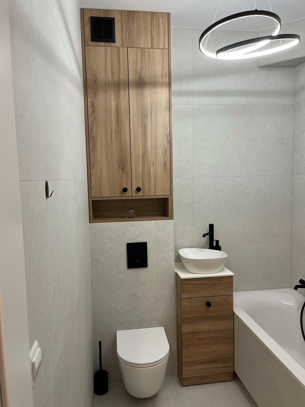 Nowoczesna łazienka z szafką na wymiar z drewnopodobnymi frontami i wiszącą umywalką nablatową oraz toaletą, oświetlona lampą w kształcie pierścieni.