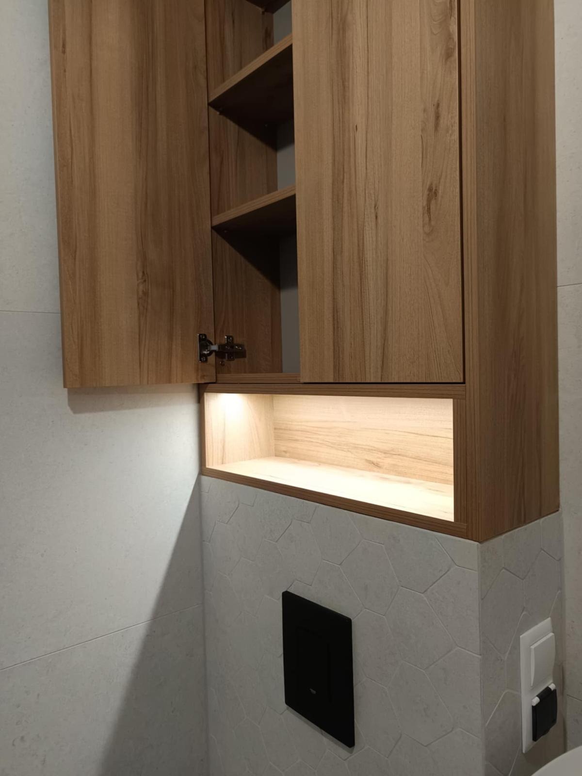 Drewniana szafka łazienkowa z otwartymi półkami i podświetleniem LED, zamontowana na ścianie z heksagonalnymi płytkami, widoczny czarny panel sterujący.