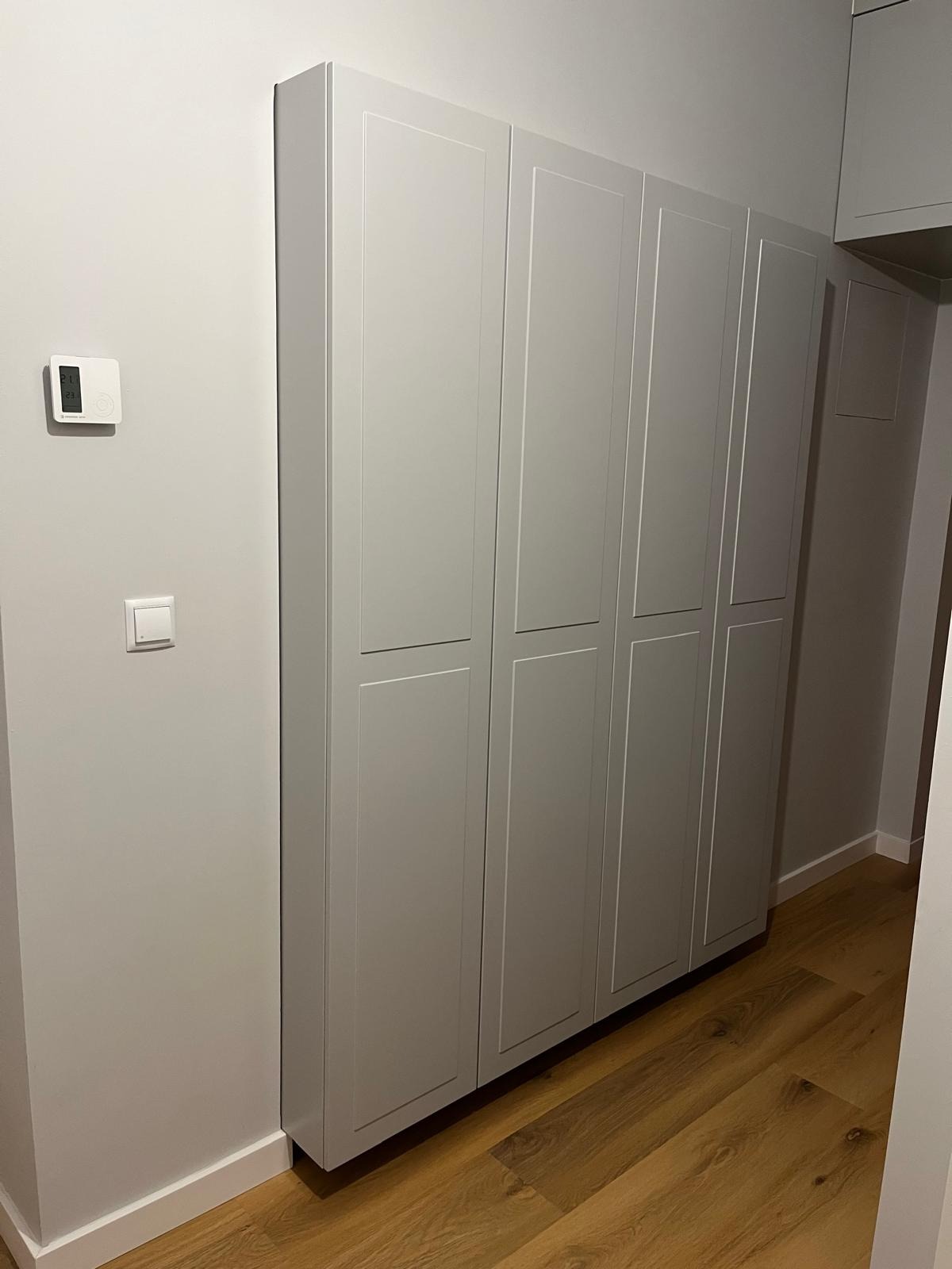 Szara szafa wnękowa z czterema segmentami drzwi w nowoczesnym przedpokoju, widoczny termostat na ścianie i drewniana podłoga.