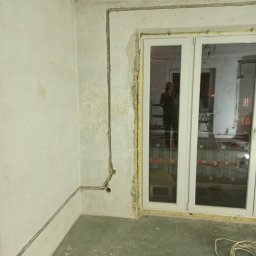 F.H.U Rafał Momro - Wewnętrzna instalacja elektryczna natynkowa w surowym pomieszczeniu z widoczną puszką instalacyjną i przewodem obok okna z odbiciem osoby w oknie.