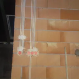 Ściana z cegły z poprowadzonymi przewodami elektrycznymi w peszlach, zamontowane puszki pod gniazdka i włączniki, widoczne pozostałości gipsu, wiadro i łopata w dolnej części kadru.