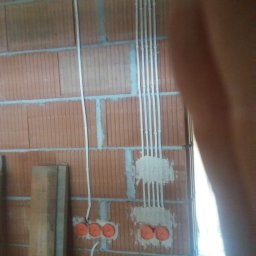 F.H.U Rafał Momro - Montaż przewodów elektrycznych w bruzdach ceglanej ściany, widoczne pomarańczowe puszki instalacyjne i pionowo ułożone peszle.