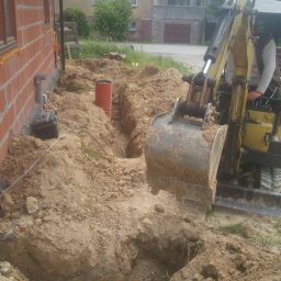 F.H.U Rafał Momro - Żółta minikoparka wykopuje rów w ziemi obok budynku z czerwonej cegły, widoczna pomarańczowa rura karbowana w wykopie.