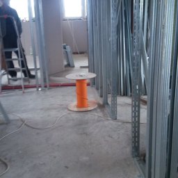 F.H.U Rafał Momro - Wnętrze w Bodzanowie w trakcie prac instalacyjnych, widoczna pomarańczowa szpula z przewodem elektrycznym, metalowe profile konstrukcyjne ścian działowych oraz pracownik na drabinie w tle.