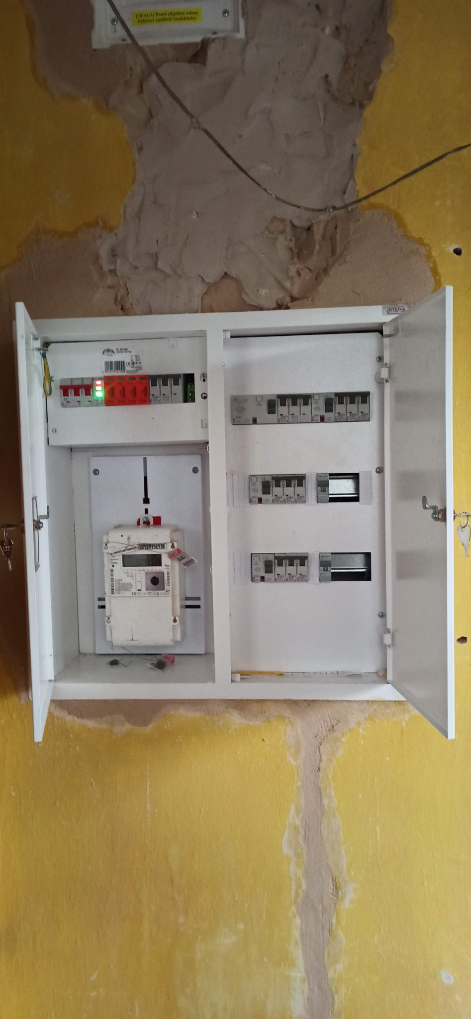 Otwarta skrzynka z bezpiecznikami, licznikiem energii elektrycznej i innymi elementami na żółtej, nierównej ścianie.