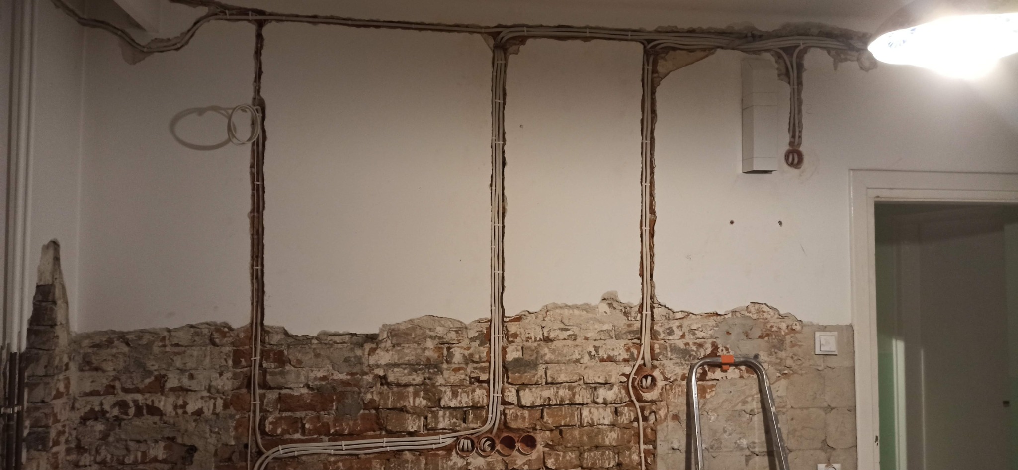 Wnętrze w trakcie remontu, widoczne bruzdy w ścianie z poprowadzonymi przewodami elektrycznymi, częściowo odsłonięta cegła, drabina oparta o ścianę, widoczny przełącznik światła i fragment drzwi.