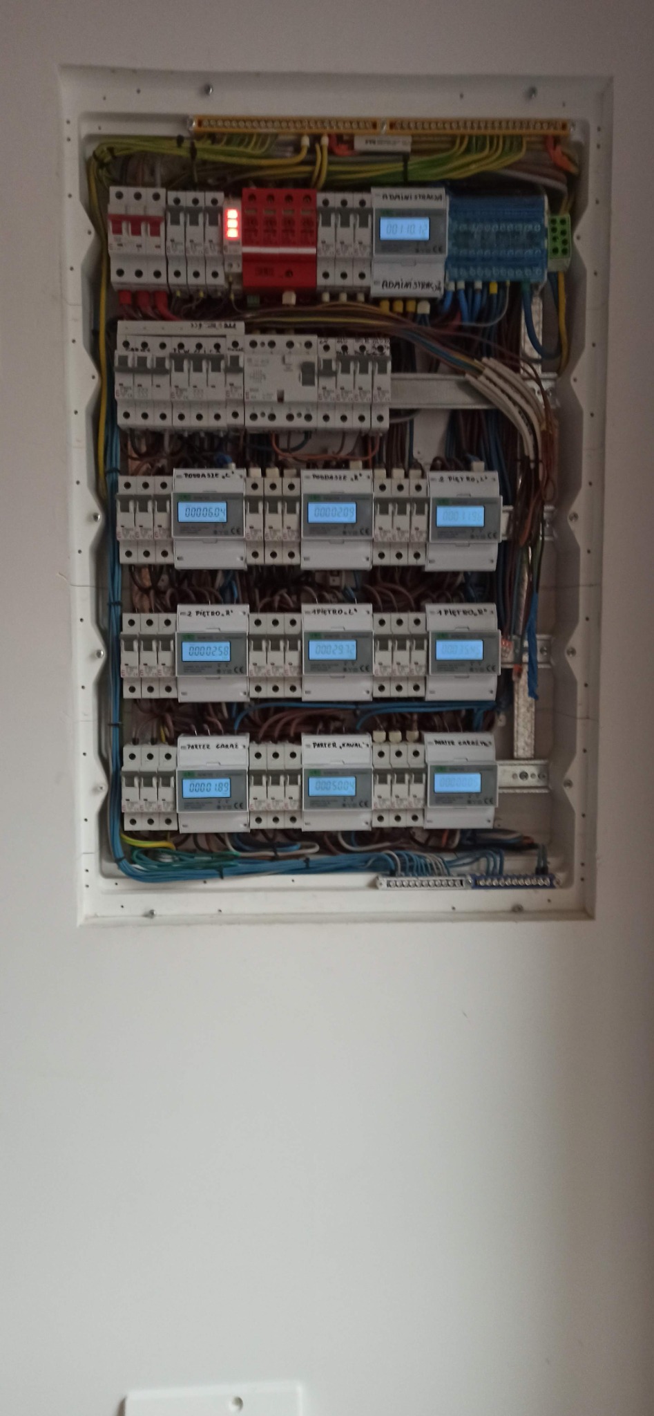 Rozdzielnica elektryczna z zamontowanymi licznikami energii, widoczne przewody, bezpieczniki i listwy zaciskowe wewnątrz białej obudowy.