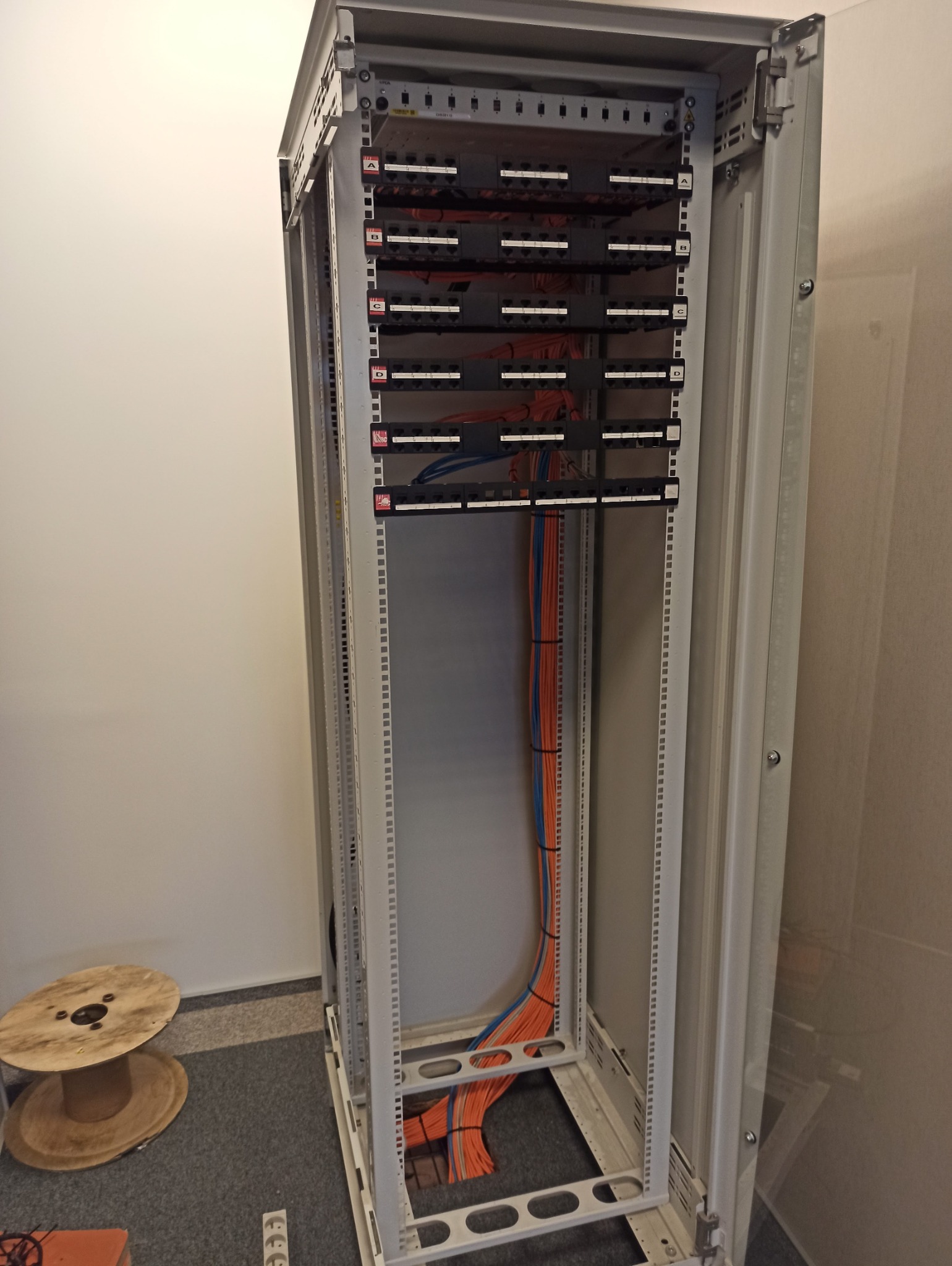 Szafa rack z panelami krosowniczymi i uporządkowanymi kablami pomarańczowymi i niebieskimi, widok z przodu, obok pusta szpula po kablach.