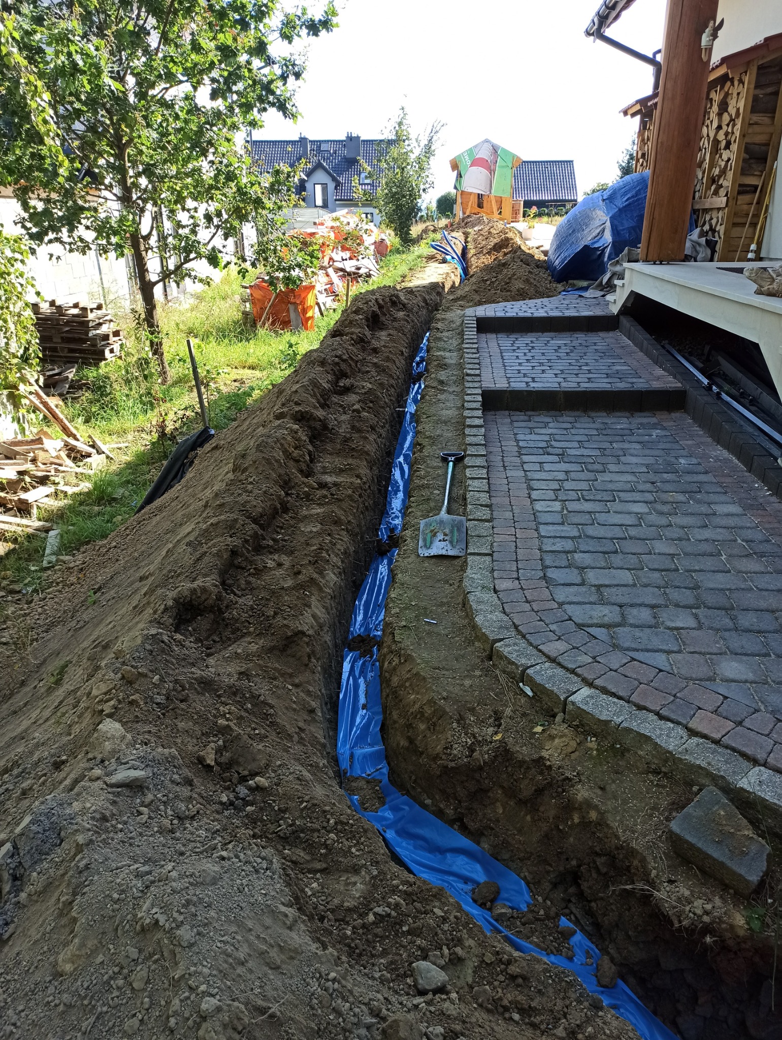 Wykop w ziemi wyłożony niebieską folią, obok kostka brukowa i łopata, widok na budynek w tle.