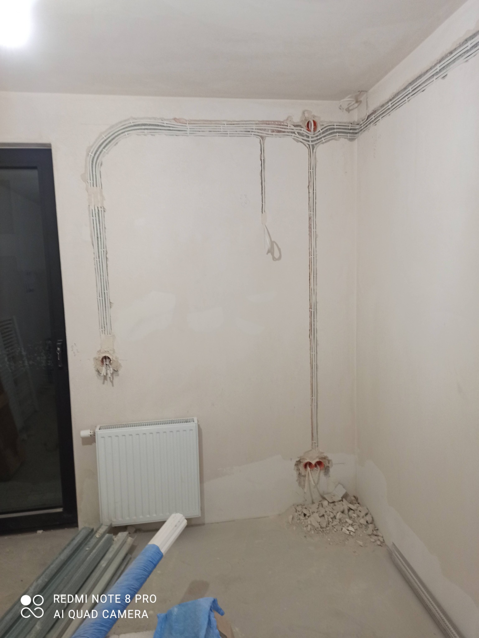 Wnętrze w trakcie remontu z widocznymi przewodami elektrycznymi w korytach na ścianach, puszkami instalacyjnymi i gruzem po kuciu.