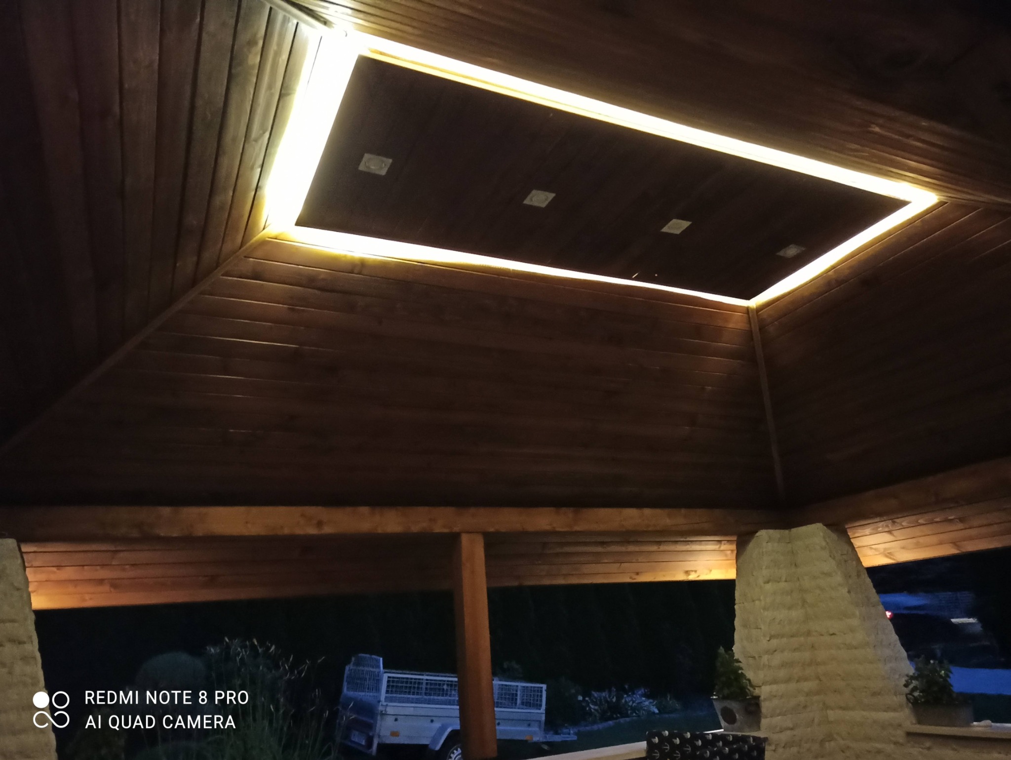 Drewniana altana z oświetleniem LED w kształcie prostokąta na suficie, widoczne reflektory punktowe, na zewnątrz przyczepka samochodowa i zieleń.