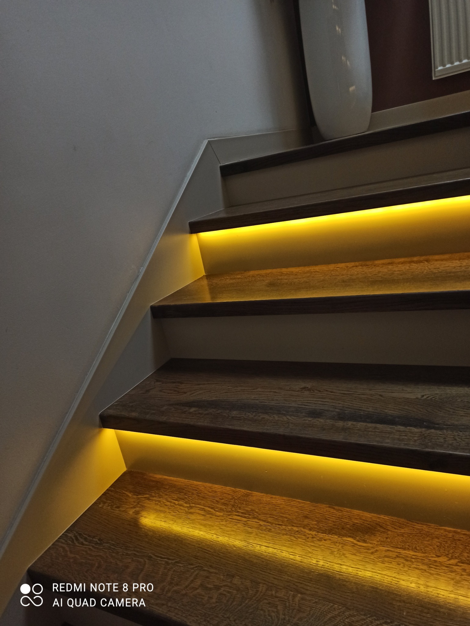 Drewniane schody z podświetleniem LED w kolorze ciepłego żółtego, oświetlające stopnie od spodu, widok z dołu, w tle ściana i fragment donicy z rośliną.