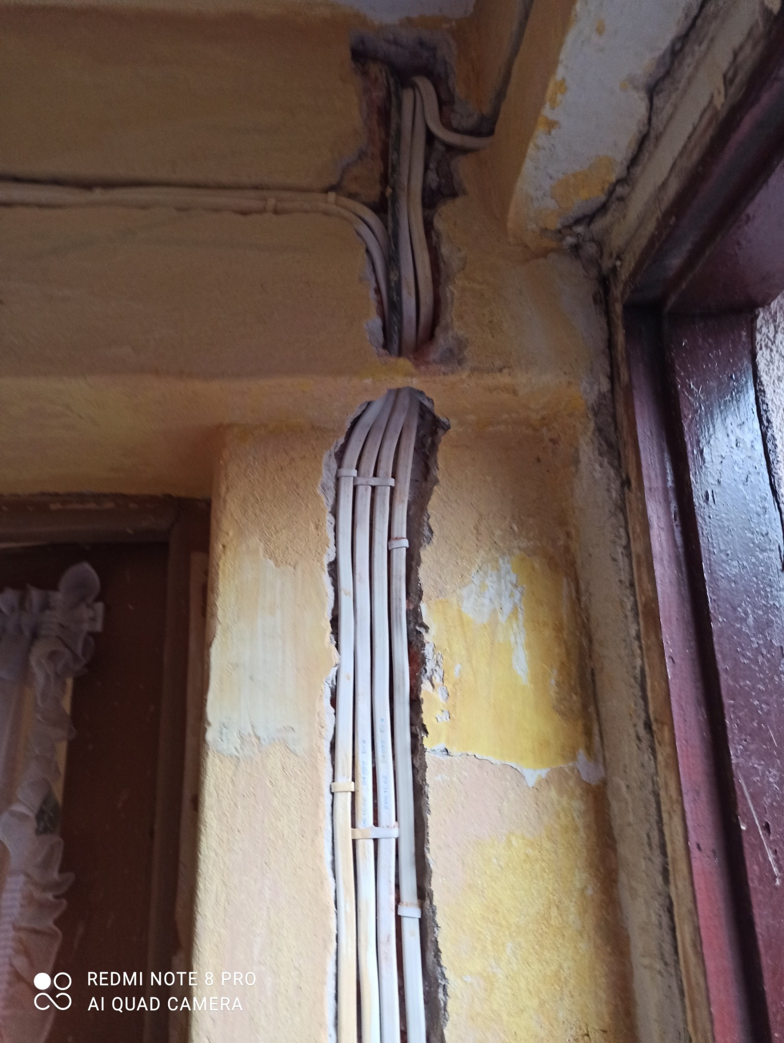 Pionowy korytarz w ścianie z widocznymi, przymocowanymi przewodami elektrycznymi, ściana w odcieniach żółci z ubytkami tynku, fragment ościeżnicy drzwi po prawej.