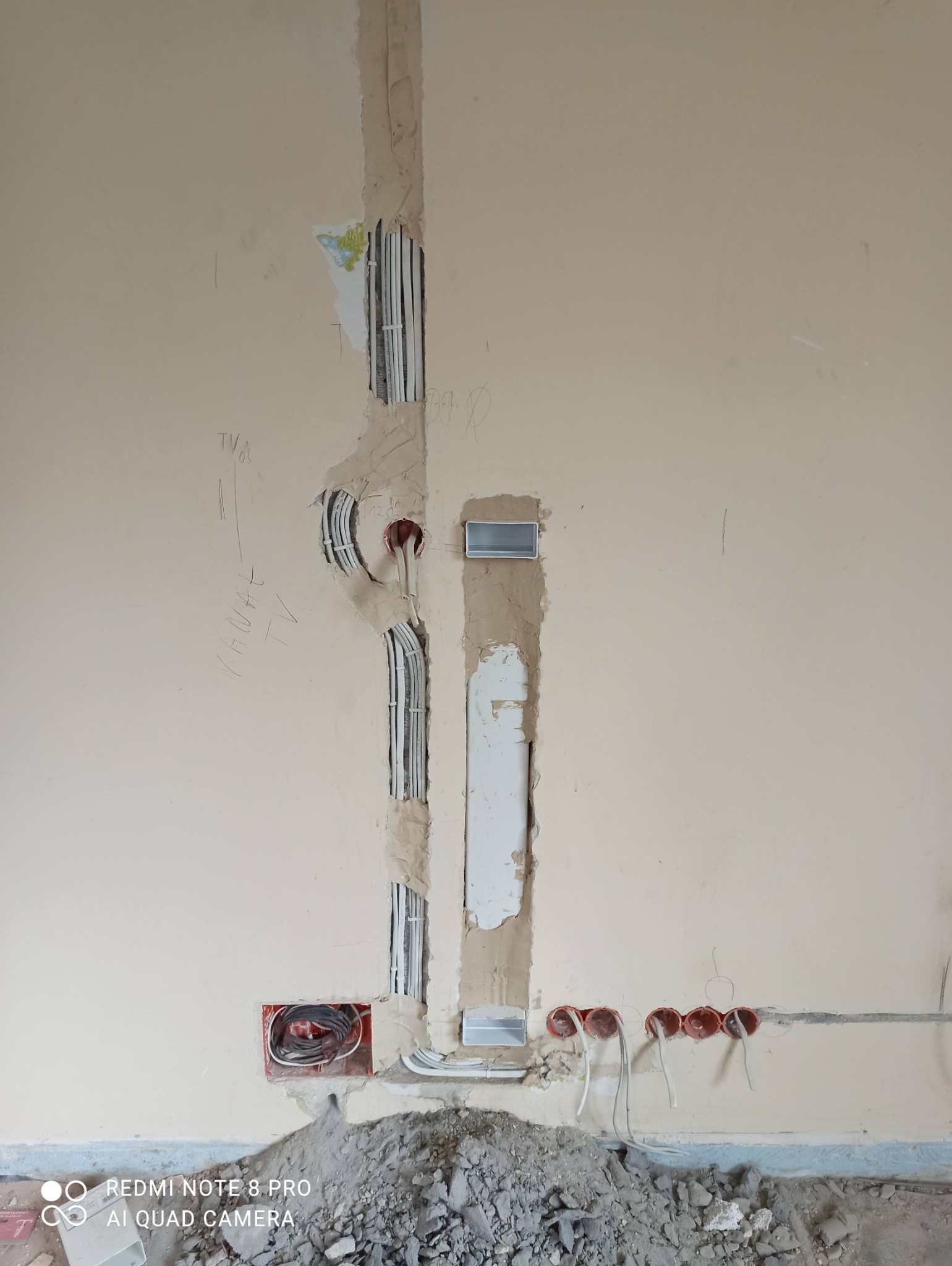 Wykute bruzdy w ścianie z poprowadzonymi kablami elektrycznymi i puszkami instalacyjnymi, widoczne ślady po kuciu i gruz na podłodze, etap prac remontowych.