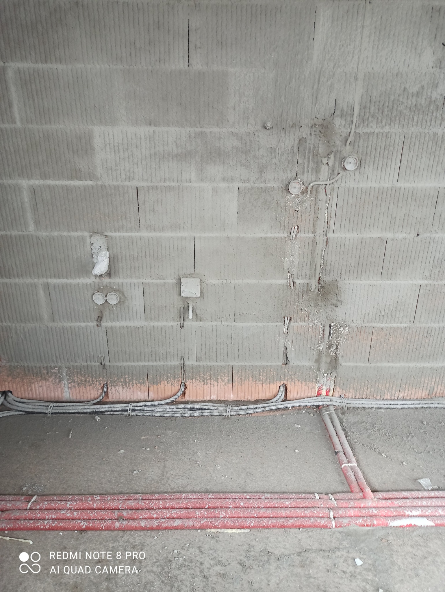 Surowy mur z widocznymi przewodami elektrycznymi w peszlach i rurach instalacyjnych w kolorze czerwonym, przygotowanymi do zalania w wylewce podłogowej.