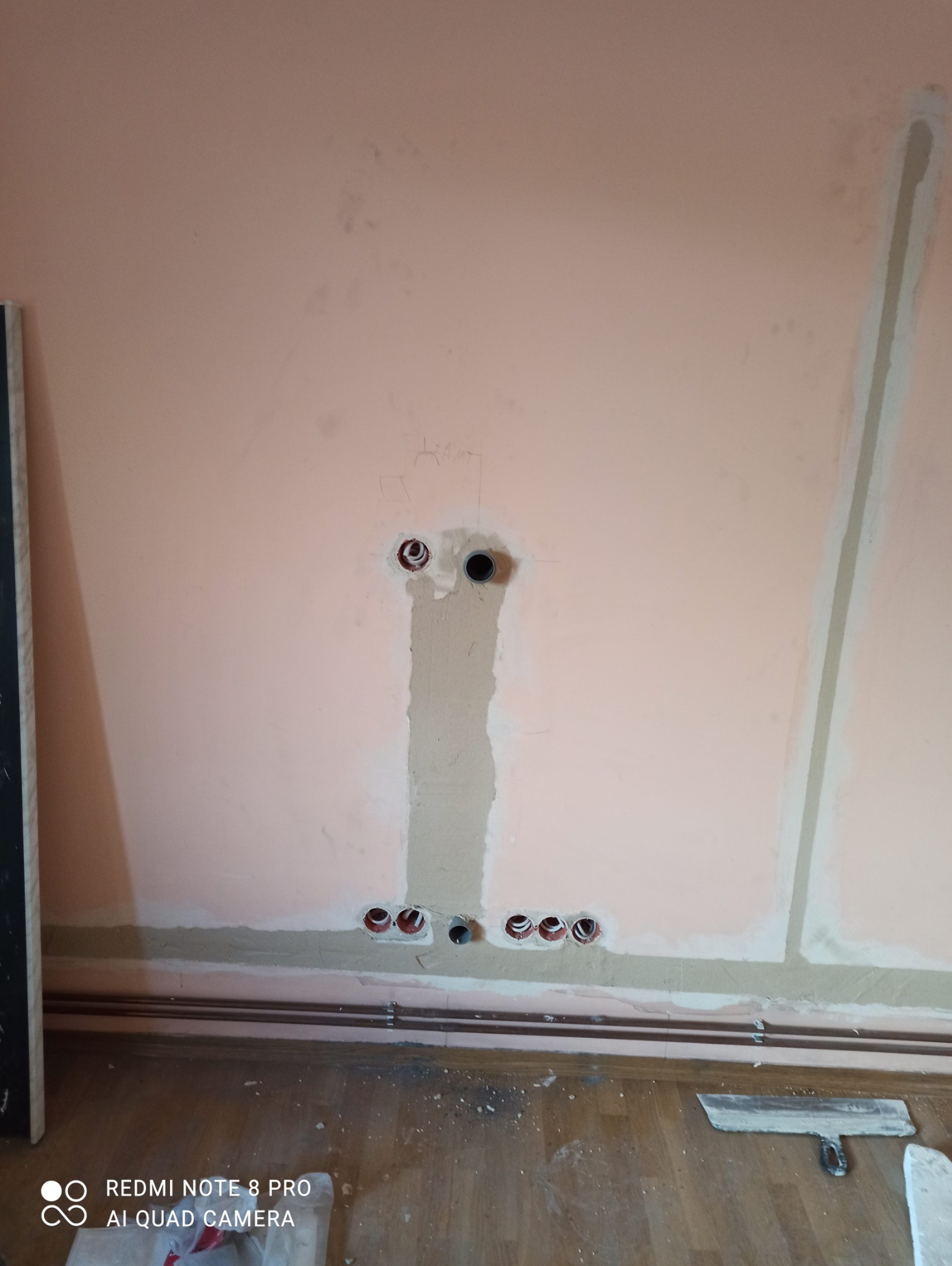 Ściana z wywierconymi otworami pod gniazdka elektryczne, częściowo zaszpachlowana, widoczne rurki instalacyjne oraz narzędzie szpachlarskie na podłodze.