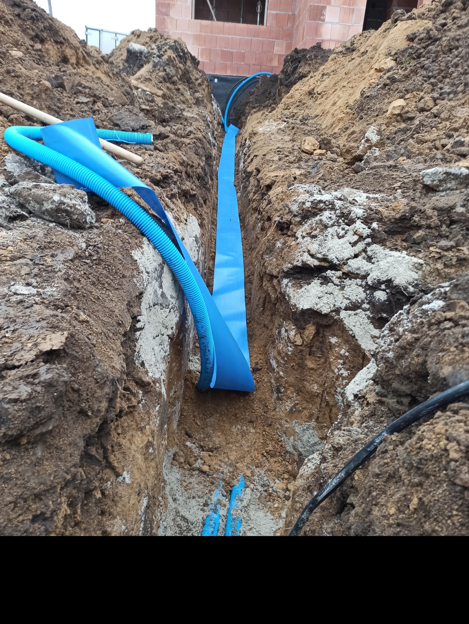 Wykop z ułożonymi niebieskimi rurami karbowanymi i płaską osłoną kablową, widoczne ściany wykopu z ziemi i fragmenty betonu, w tle mur z czerwonej cegły.