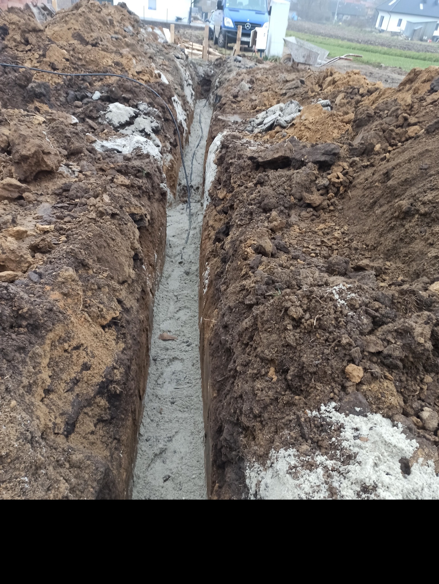 Wykop w ziemi wypełniony betonem, widoczne kable elektryczne wewnątrz, gruz i ziemia po bokach, w tle samochód dostawczy i domy.
