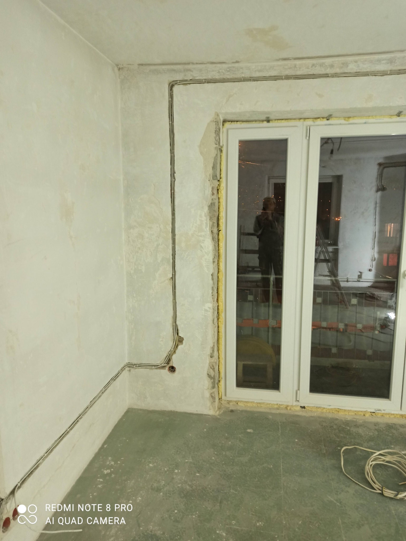 Wewnętrzna instalacja elektryczna natynkowa w surowym pomieszczeniu z widoczną puszką instalacyjną i przewodem obok okna z odbiciem osoby w oknie.