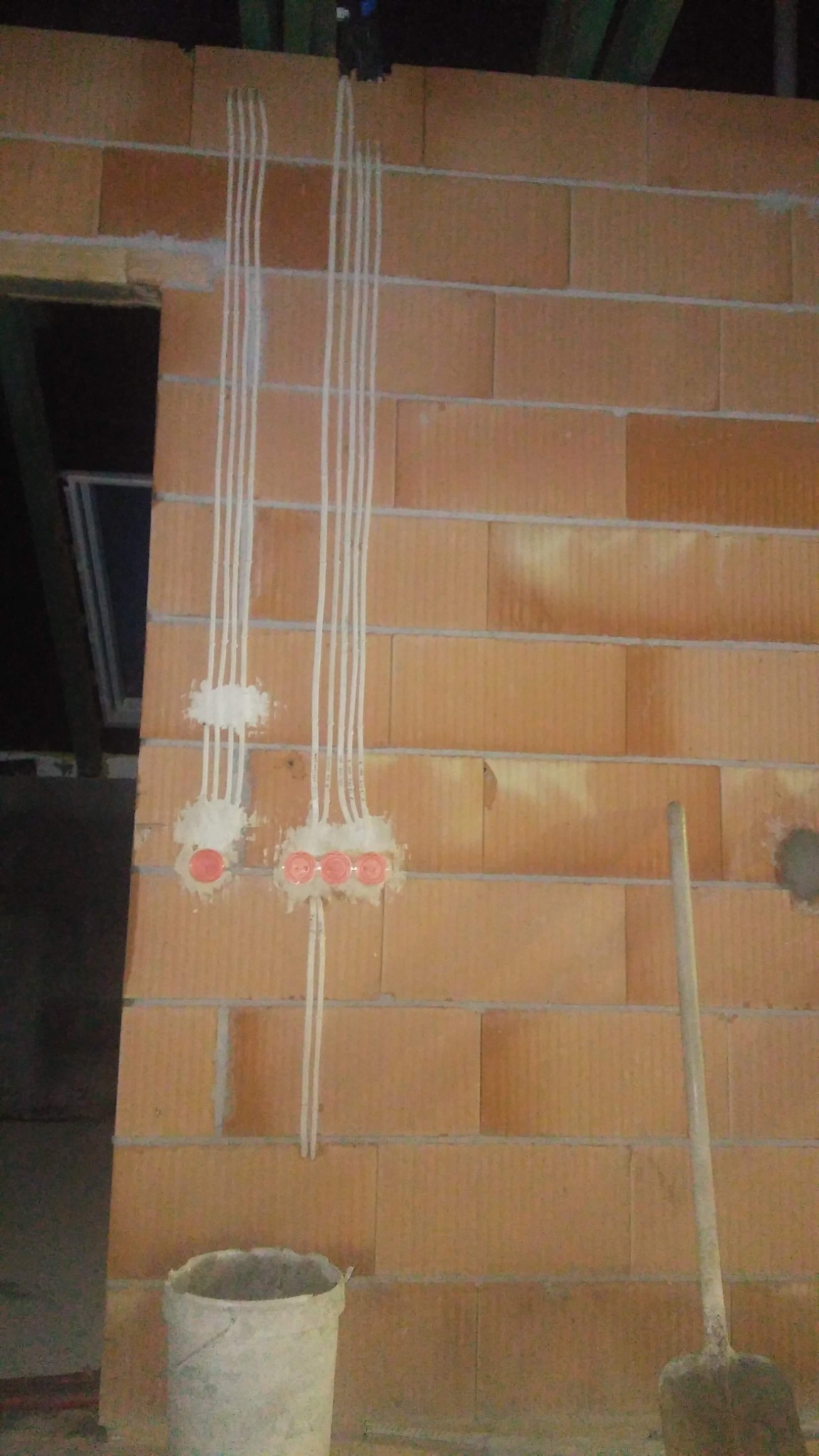 Ściana z cegły z poprowadzonymi przewodami elektrycznymi w peszlach, zamontowane puszki pod gniazdka i włączniki, widoczne pozostałości gipsu, wiadro i łopata w dolnej części kadru.