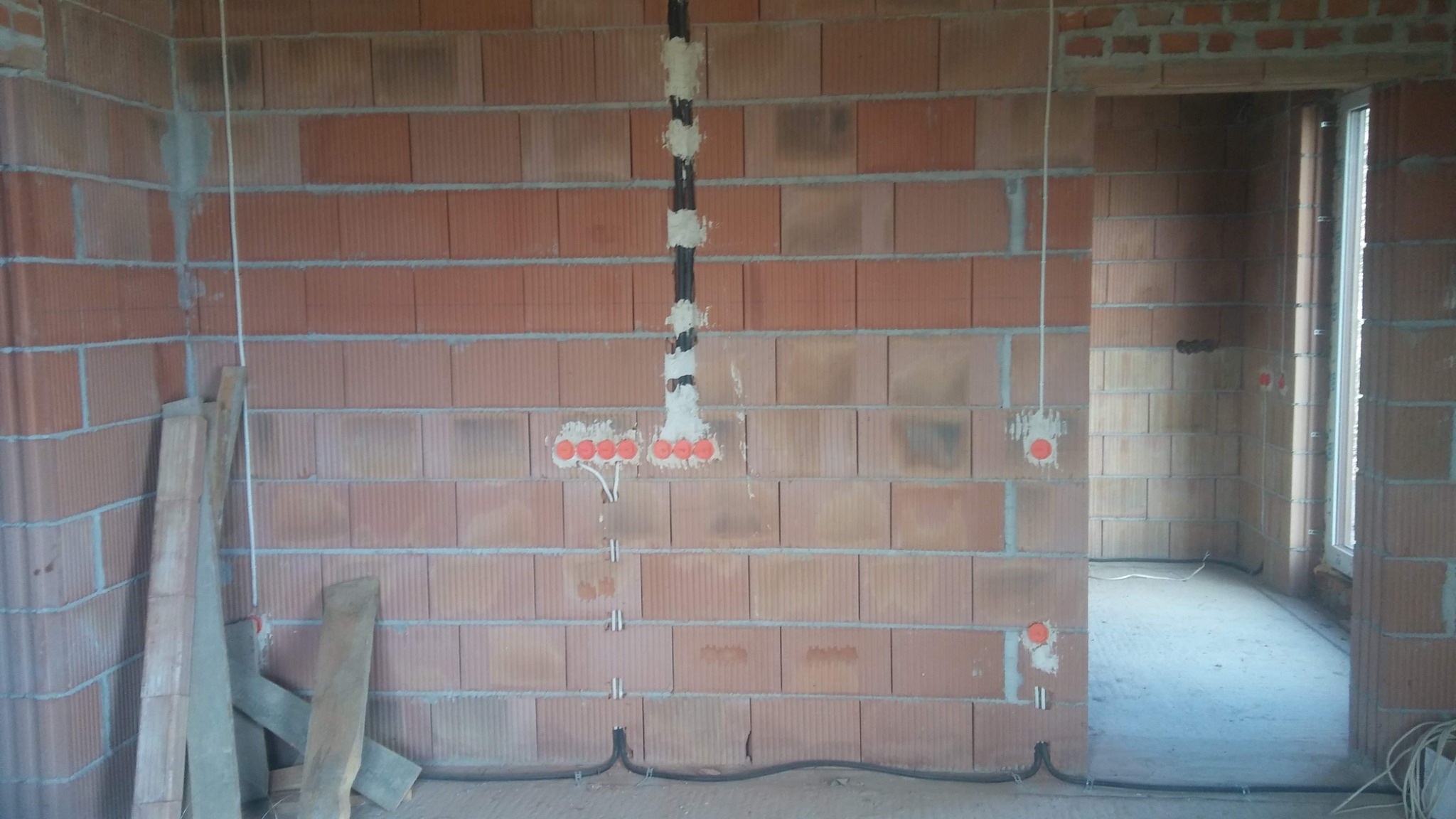 Wnętrze w trakcie prac elektrycznych: kable prowadzone po ścianie z czerwonej cegły, puszki pod gniazdka i włączniki zalane gipsem, widoczne przewody pod podłogą.