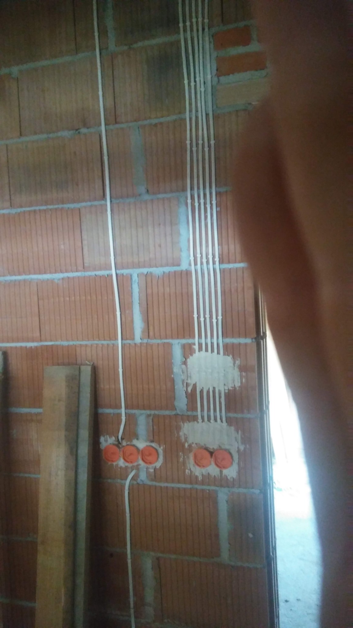 Montaż przewodów elektrycznych w bruzdach ceglanej ściany, widoczne pomarańczowe puszki instalacyjne i pionowo ułożone peszle.
