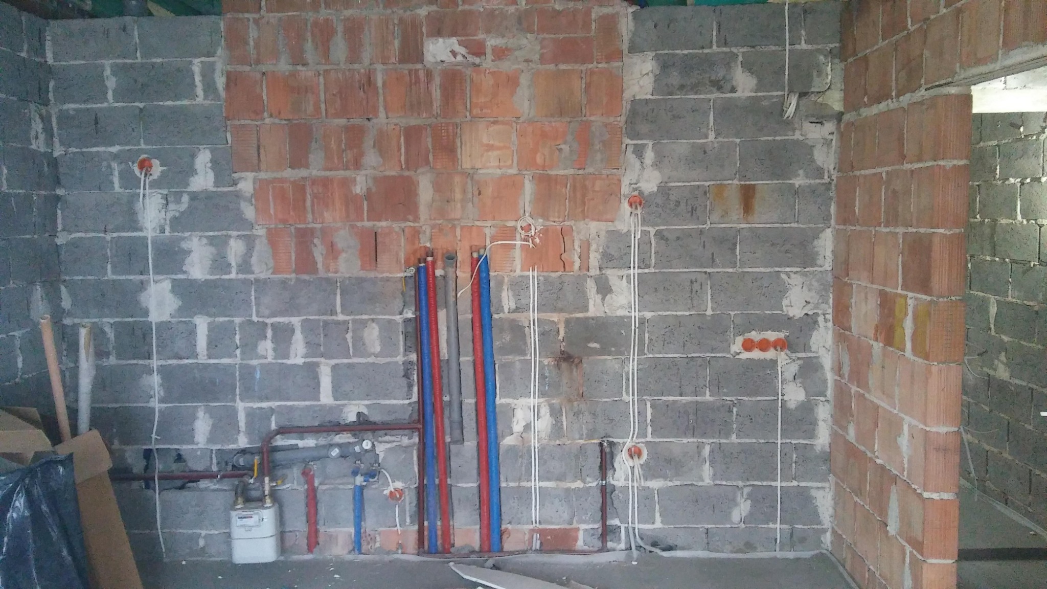 Surowa ściana z bloków i cegieł z widocznymi przewodami elektrycznymi w peszlach, puszkami instalacyjnymi i rurami instalacji wodnej oraz gazowej w trakcie montażu.