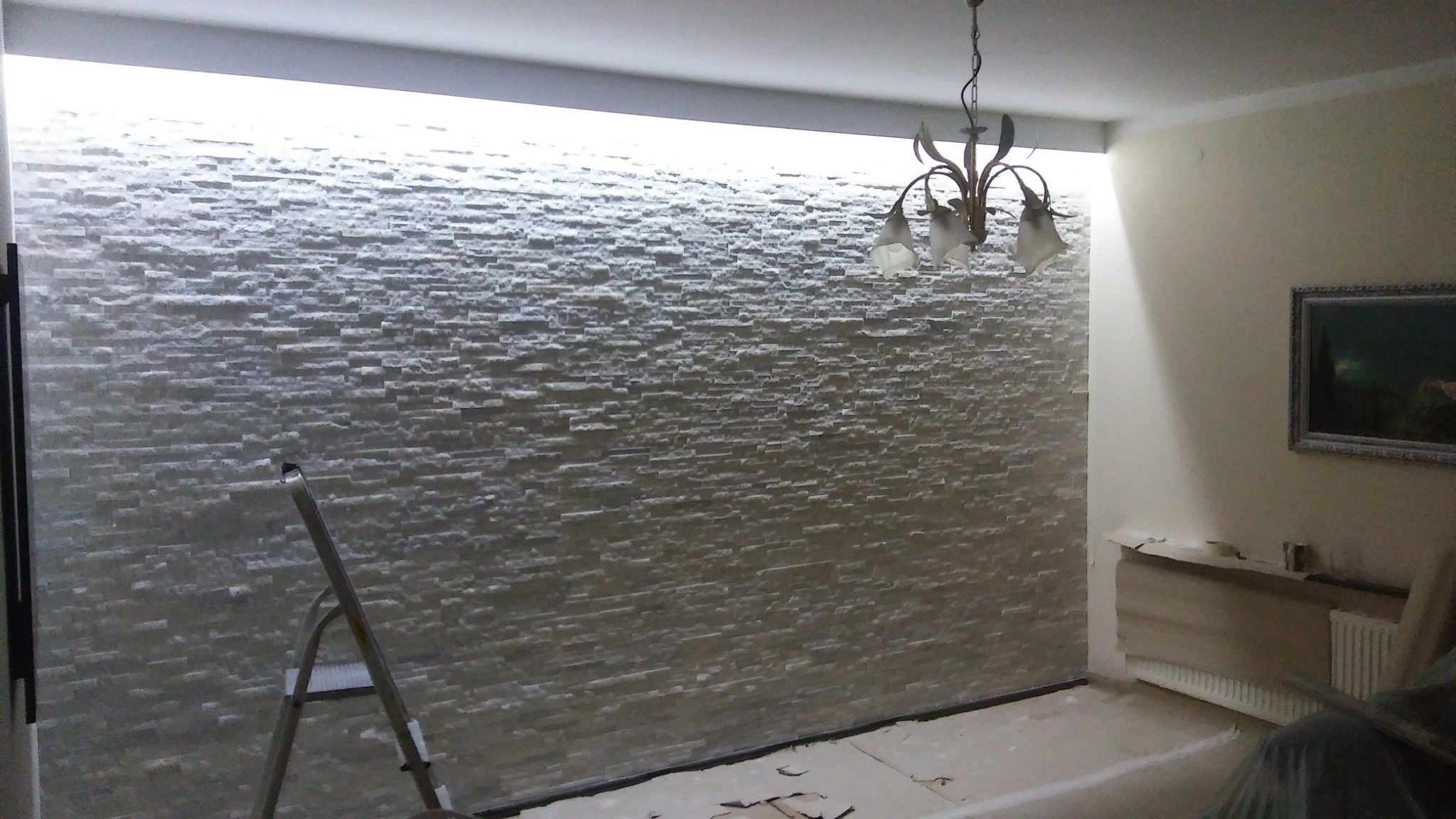 Ściana wyłożona dekoracyjnym kamieniem w pomieszczeniu z oświetleniem LED podsufitowym, widoczna drabina i lampa wisząca.