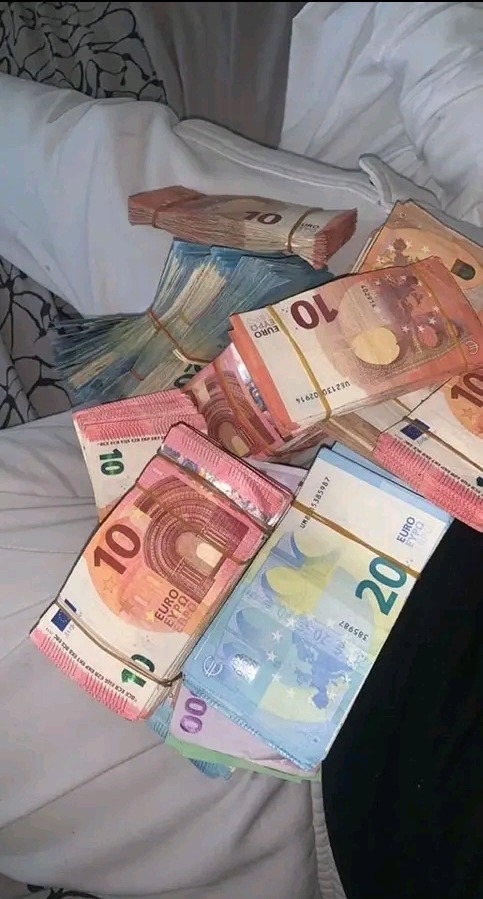Stosy banknotów euro różnych nominałów (10, 20, 100) związanych gumkami, ułożone na białej pościeli, z fragmentem czarnego materiału w rogu.